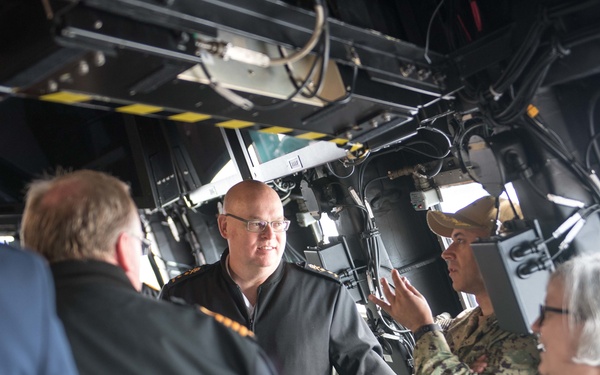 A group of individuals, including at least one person in US Navy camouflage uniform and others in dark naval or civilian attire, are gathered inside what appears to be a military vessel's bridge or command area. A uniformed service member appears to be briefing or explaining something to the group, gesturing with their hand. The setting is characterized by overhead equipment, cables, and yellow hazard markings typical of a warship interior.