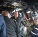 A group of individuals, including at least one person in US Navy camouflage uniform and others in dark naval or civilian attire, are gathered inside what appears to be a military vessel's bridge or command area. A uniformed service member appears to be briefing or explaining something to the group, gesturing with their hand. The setting is characterized by overhead equipment, cables, and yellow hazard markings typical of a warship interior.