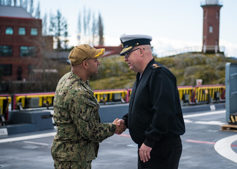 Two military personnel are shaking hands on what appears to be a ship's flight deck or pier. One individual is wearing U.S. Navy NWU Type III camouflage uniform with a tan cap, while the other is wearing a dark naval dress uniform with a white-topped officer's cap and gold insignia. The setting appears to be a port or naval facility with urban infrastructure visible in the background.