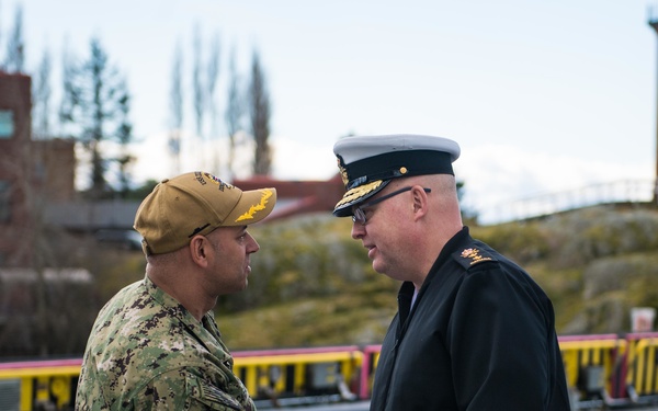 Two military personnel are shaking hands on what appears to be a ship's flight deck or pier. One individual is wearing U.S. Navy NWU Type III camouflage uniform with a tan cap, while the other is wearing a dark naval dress uniform with a white-topped officer's cap and gold insignia. The setting appears to be a port or naval facility with urban infrastructure visible in the background.
