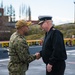 Two military personnel are shaking hands on what appears to be a ship's flight deck or pier. One individual is wearing U.S. Navy NWU Type III camouflage uniform with a tan cap, while the other is wearing a dark naval dress uniform with a white-topped officer's cap and gold insignia. The setting appears to be a port or naval facility with urban infrastructure visible in the background.