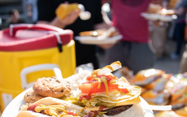 A person holds a white paper plate loaded with a hot dog, a burger with toppings, and a cookie at what appears to be an outdoor cookout or tailgate event. The background shows a food serving table with chip bags, additional food items, and a large yellow and red beverage cooler. Multiple people are visible in the blurred background, suggesting a social gathering.