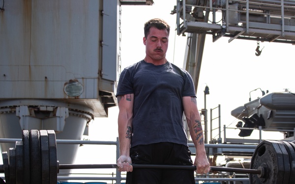 A man with tattoos and a mustache is performing a deadlift with a heavily loaded barbell on what appears to be the deck of a naval vessel. Two other men are visible nearby — one wearing a Billabong t-shirt observing from the left, and another seated on the right watching. A container of gym chalk is visible on the deck, indicating a structured workout session.