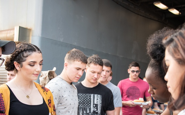 A group of young people are lined up at an outdoor food serving station, receiving food from servers wearing blue disposable gloves. The setting appears to be a casual outdoor cookout or barbecue event, with foil trays of food visible on the serving table. The atmosphere is informal and social, suggesting a community or group gathering.