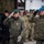20th anniversary of NATO expansion Two senior military officers in the foreground are rendering salutes during what appears to be a formal military ceremony. The officer on the left wears a green beret and olive-colored uniform, while the officer on the right wears a blue beret and camouflage uniform with a Polish flag patch. A formation of military personnel from multiple nations is visible in the background, suggesting a multinational military event.