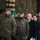 20th anniversary of NATO expansion A formal military ceremony taking place outdoors in cold, lightly snowy weather. Two uniformed military officers in the foreground are rendering salutes, accompanied by civilian dignitaries standing at attention. The setting appears to be a Polish military event, evidenced by Polish flag patches on uniforms and Polish text visible on a sign.