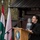 20th anniversary of NATO expansion A woman in black attire is speaking at a podium bearing the emblem of the XVI Polish Military Contingent EUFOR ALTHEA during an official ceremony. The setting appears to be an outdoor military event with multiple national flags visible in the background, including Hungarian and Czech flags, along with a sign reading 'POLAND'. Display boards referencing NATO and Poland are visible behind her.