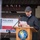 20th anniversary of NATO expansion A man in a grey jacket and plaid flat cap is delivering a speech at a podium during an official ceremony. The podium bears the emblem of the Polish Military Contingent EUFOR ALTHEA, and a banner in the background reads 'Poland - 20 Years in NATO'. The setting appears to be an outdoor or semi-covered ceremonial event.