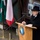 20th anniversary of NATO expansion A man in a black coat and hat is speaking at a podium during an official ceremony. The podium bears the insignia of the Polish Military Contingent EUFOR ALTHEA, XVI rotation. The setting appears to be an outdoor or semi-covered official event related to Poland's participation in NATO and EU military operations.