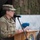 20th anniversary of NATO expansion A female U.S. military officer wearing a camouflage patrol cap and olive drab fleece jacket is speaking at a podium with a microphone. She appears to be reading from papers at what looks like an official military ceremony or event. A wooden lectern bearing a military unit insignia is visible in the foreground, with other military personnel visible in the blurred background.