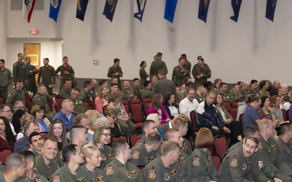 A military ceremony or graduation event is taking place in a large indoor hall. A group of uniformed personnel wearing olive green flight suits with patches are seated in the foreground, while civilians and additional military personnel fill the background. Flags and pennants are displayed along the upper walls of the venue.