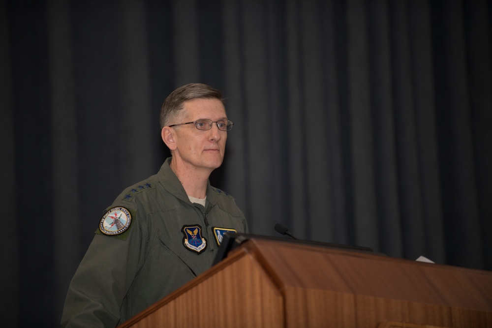 A military officer in an olive green flight suit stands at a wooden podium, appearing to deliver a speech or address. He wears glasses and has multiple patches visible on his left sleeve and chest, indicating a senior rank. The setting appears to be a formal auditorium or conference room with dark curtain backdrop.