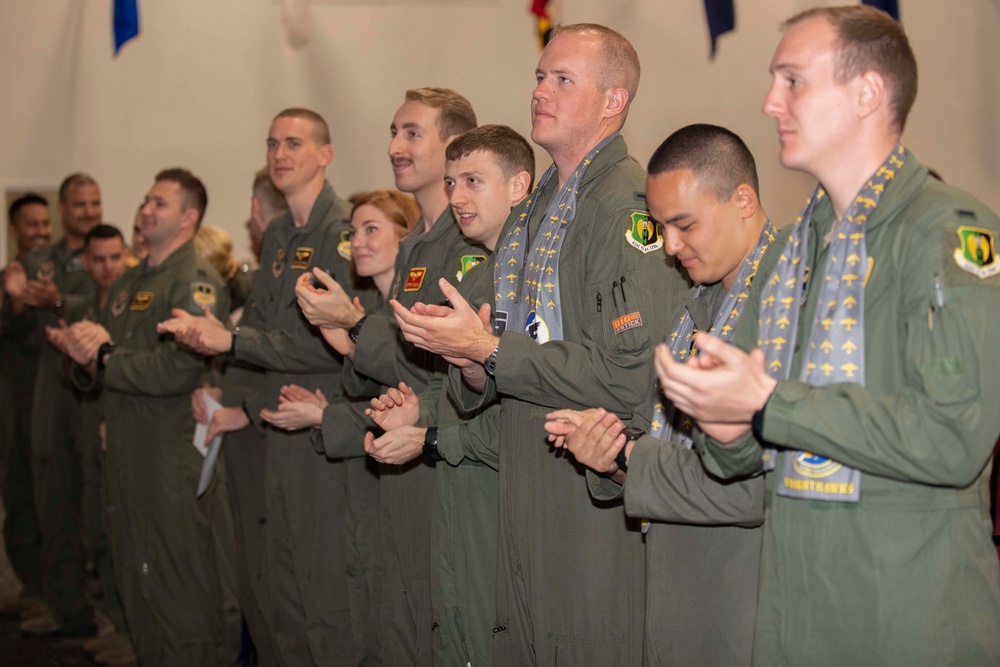 A group of military personnel dressed in olive green flight suits are standing in a line and applauding at what appears to be a formal ceremony or graduation event. Several individuals are wearing decorative blue scarves with gold aircraft motifs, suggesting a pilot training graduation or wings ceremony. The group includes both men and women, and unit patches are visible on their flight suits.