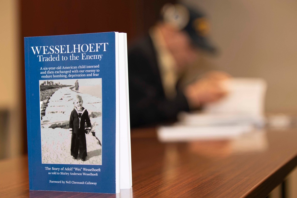 A blue hardcover book titled 'Wesselhoeft: Traded to the Enemy' is displayed upright on a wooden table in sharp focus. The book tells the story of Adolf 'Wes' Wesselhoeft, a six-year-old American child interned and exchanged with the enemy during wartime. In the blurred background, a person wearing a military-style cap appears to be seated at the same table.