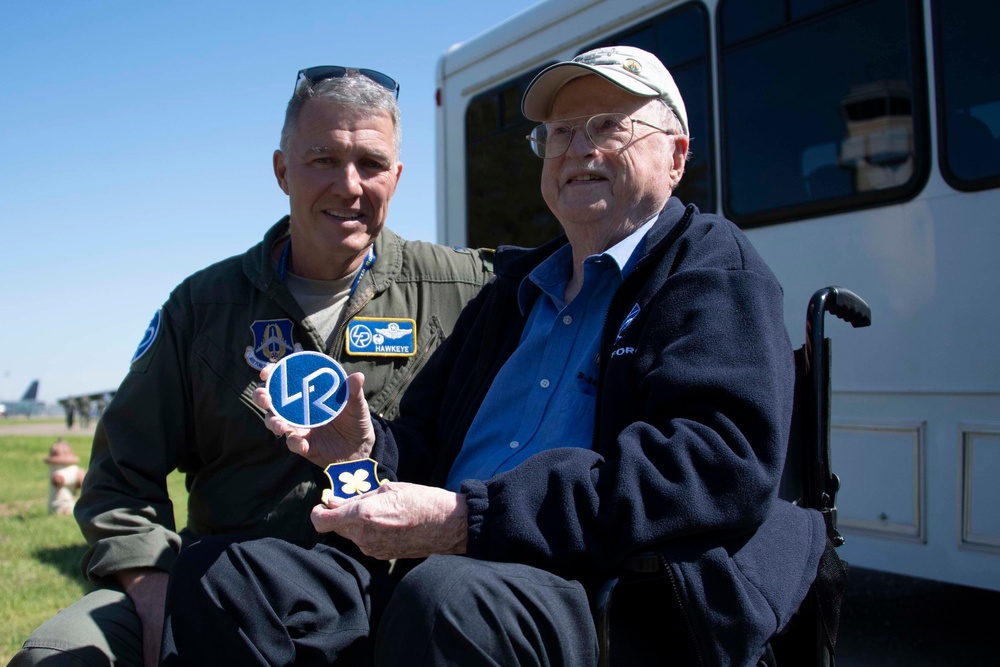 Two men pose together outdoors near a white bus on what appears to be an airfield. A military aviator in an olive flight suit with patches, including one labeled 'HAWKEYE,' holds a circular blue-and-white patch with 'LR' lettering, while an elderly man in a wheelchair holds a shield-shaped military patch. The setting and attire suggest a ceremonial or commemorative aviation event.