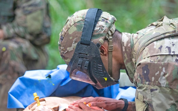 A military soldier in MultiCam/OCP camouflage uniform and combat helmet is performing medical training on a realistic human mannequin/training dummy placed on a litter/stretcher. The scene appears to be an outdoor tactical combat casualty care (TCCC) training exercise, with a blue medical drape visible beneath the mannequin. At least one additional soldier is partially visible in the background.