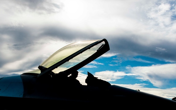 A fighter jet is photographed in dramatic silhouette on an airfield tarmac, with its canopy open and a pilot or crew member visible in the cockpit. The image is backlit by a partially cloudy sky with the sun breaking through, creating strong contrast between the dark aircraft and the bright sky. The low-angle perspective emphasizes the sleek, aerodynamic profile of the aircraft.
