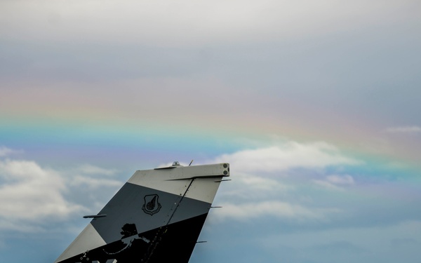 The image shows the tail fin of a U.S. Air Force fighter aircraft, marked with 'AK' and '18 AGRS' designations, indicating it belongs to the 18th Aggressor Squadron based in Alaska. A rainbow is visible in the partly cloudy sky in the background. The aircraft tail features a black and gray paint scheme with a unit insignia patch and a map of Alaska.