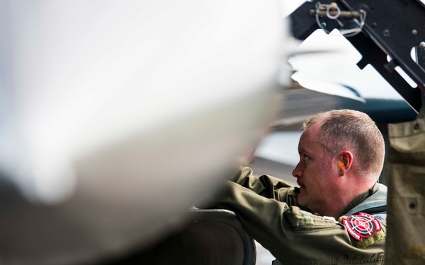 A military aviator or ground crew member in flight gear is conducting a close inspection of a military aircraft, likely performing a pre-flight or post-flight check. The individual is wearing a green flight suit with patches, survival vest, and ear protection. The image is taken from a tight angle near the aircraft's engine intake or fuselage.