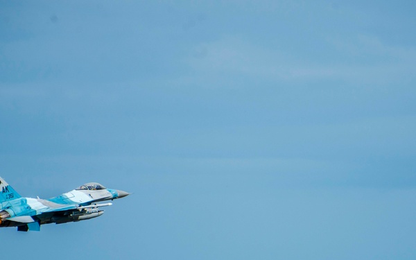 A military fighter jet, appearing to be an F-16 Fighting Falcon painted in a light blue aggressor scheme, is captured in low-level flight just above a treeline. The aircraft displays 'AK' tail codes, indicating it belongs to an Alaska-based unit, likely the 18th Aggressor Squadron from Eielson Air Force Base. The jet appears to be in a climbing or level flight attitude with afterburner engaged, visible from the engine exhaust glow.