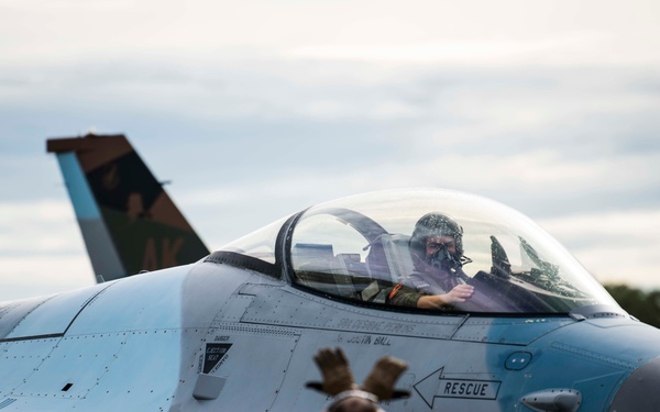 A military F-16 Fighting Falcon jet aircraft, numbered 351, is on a flight line tarmac with a pilot seated in the cockpit wearing a flight mask and helmet. Ground crew members in high-visibility yellow vests are positioned in front of the aircraft, appearing to conduct pre-flight or post-flight marshaling procedures. A second aircraft with an 'AK' tail marking is partially visible in the background.