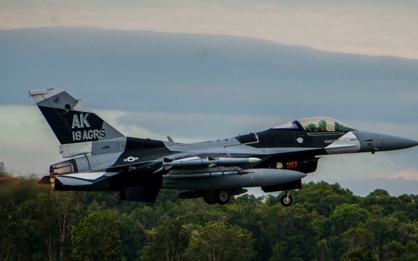 A U.S. Air Force F-16 Fighting Falcon is captured just above the runway during takeoff, with afterburner visibly engaged. The aircraft bears tail markings 'AK' and '18 AGRS', identifying it as belonging to the 18th Aggressor Squadron based in Alaska. The image was taken at what appears to be a military airfield surrounded by dense tree lines under overcast skies.