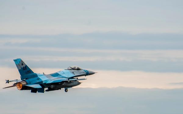 A U.S. Air Force F-16 Fighting Falcon with 'AK' tail code markings is captured just after takeoff or during a low-altitude pass over a runway. The aircraft is painted in a distinctive blue-gray aggressor scheme and appears to be carrying air-to-air missiles on its wingtips. A vehicle and runway marker are visible in the background near a treeline.