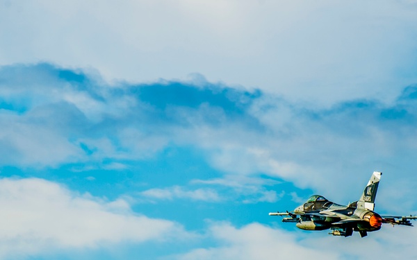 A military fighter jet, consistent with an F-16 Fighting Falcon, is captured in flight against a partly cloudy blue sky. The aircraft appears to be in level flight or a slight climb, with its afterburner visibly engaged, producing an orange glow at the engine exhaust. The image is sharp and well-composed, with the jet positioned in the right-center of the frame.