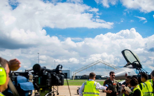 A media press event is taking place on a military airfield tarmac, where a pilot in a flight suit is being interviewed by journalists and media personnel. Several individuals wearing high-visibility yellow vests labeled 'AIR FORCE' are present, along with camera operators filming the scene. A military jet aircraft, likely an F-16 or similar fighter jet, is visible in the background.