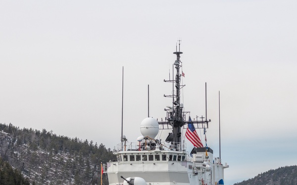 A U.S. Coast Guard cutter bearing hull number 906 is underway in a cold-weather coastal waterway surrounded by snow-dusted, forested hills. The vessel displays the distinctive USCG diagonal red stripe on its bow and flies the American flag from its mast. The overcast sky and icy shoreline suggest a winter or early spring setting in a northern region.