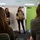 A woman is presenting to a small group in what appears to be a classroom or training room setting, holding up a military-style camouflage body armor vest. A whiteboard behind her reads 'Group 2 Survival Vests / Body Armor,' indicating this is a group presentation or training exercise. Two audience members are visible from behind in the foreground, and a second presenter stands nearby.