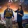 A woman in a blue checkered shirt is presenting or demonstrating a tactical/military-style vest worn by another woman in a blue shirt and gray pants. The presentation appears to be taking place in front of a large space-themed backdrop with the partial text 'AFWERX' visible, suggesting a U.S. Air Force innovation or technology event. An audience is partially visible in the foreground.