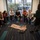 A group of approximately seven women are gathered in a modern office conference room, seated in a semicircle facing a presenter who is standing and speaking. A garment (appears to be military-style pants or uniform) is laid out on the floor in the center of the group, and military boots are visible on a table to the right. The setting suggests a professional training, workshop, or briefing session.