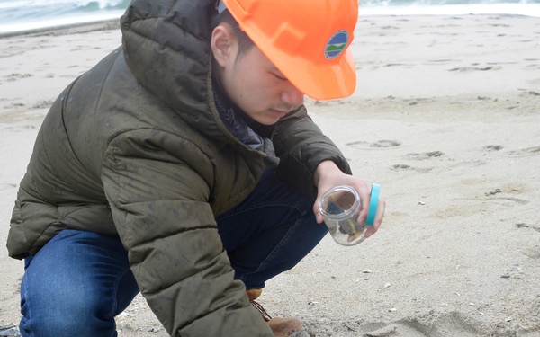 A person wearing an orange hard hat, olive puffer jacket, jeans, and purple nitrile gloves is crouched on a sandy beach collecting samples. They are holding a small glass jar with a teal lid and appear to be gathering dark, tar-like or oily debris mixed with organic material from the sand. A yellow reference card or data sheet is visible on the sand nearby, suggesting a scientific or environmental assessment activity.