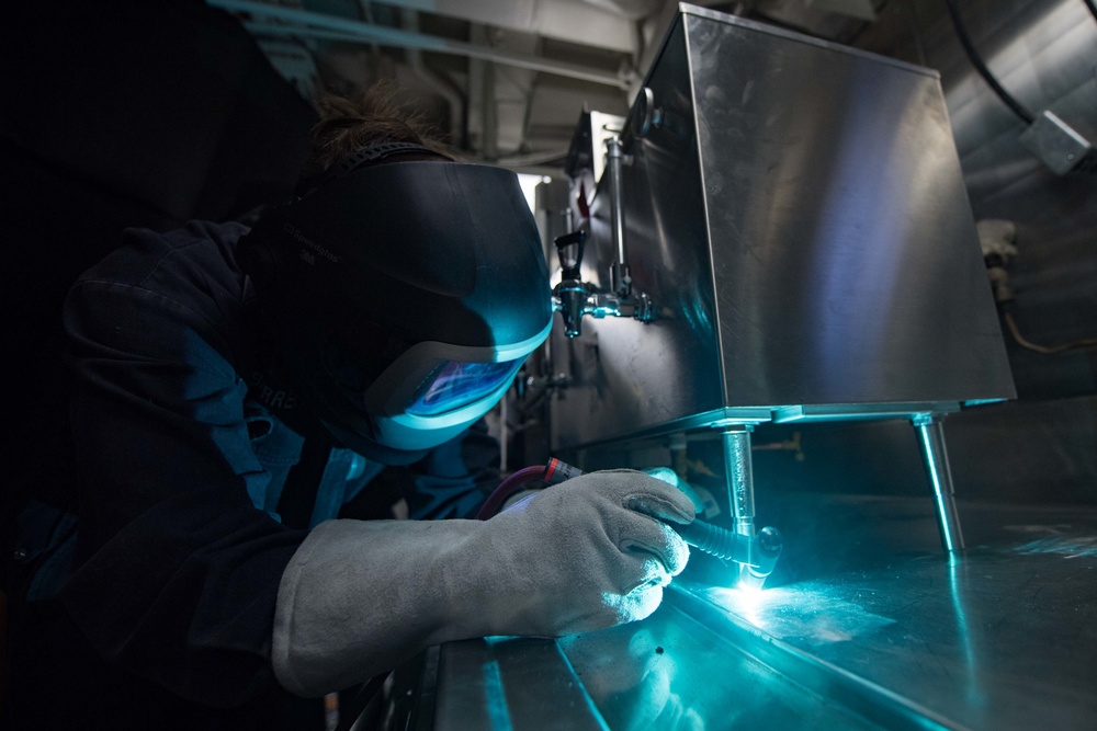 U.S. Navy Hull Maintenance Technician welds a coffee machine to a table aboard USS Stockdale