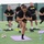 A group of U.S. Army personnel in matching black 'ARMY' PT (physical training) uniforms are performing yoga or stretching exercises on purple yoga mats on an artificial turf field. A female soldier in the foreground is in a balancing pose with hands pressed together in a prayer position. The setting appears to be an outdoor sports facility, likely on a military installation.