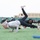 A large group of people are performing yoga or fitness exercises on a grass field, likely at a military installation based on the 'ARMY' text visible on some clothing. The participants are in various plank and leg-raise positions on yoga mats and directly on the turf. An older individual in a gray hoodie is prominently featured in the foreground alongside others in black athletic wear.