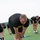 A group of individuals wearing U.S. Army physical training uniforms (black shirts with yellow 'ARMY' lettering and black shorts) are performing stretching exercises on a grass field. The foreground subject is barefoot, standing on a blue towel, and bending forward to touch his shins in a hamstring stretch. Multiple other soldiers are visible in the background performing similar stretching movements.