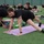 A large group of U.S. Army personnel in matching black 'ARMY' t-shirts and shorts are participating in an outdoor fitness or yoga session on an artificial turf field. A smiling woman in the foreground is performing a stretching exercise on a pink yoga mat with text written on it. The session appears organized and involves multiple participants of varying ages performing similar movements.