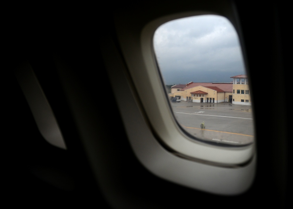 A view through an airplane window showing an airport tarmac on a cloudy, overcast day. The wet tarmac suggests recent rainfall, and a terminal or hangar building with a yellow facade and red roof is visible in the background. The aircraft appears to be stationary at the airport.