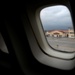 A view through an airplane window showing an airport tarmac on a cloudy, overcast day. The wet tarmac suggests recent rainfall, and a terminal or hangar building with a yellow facade and red roof is visible in the background. The aircraft appears to be stationary at the airport.