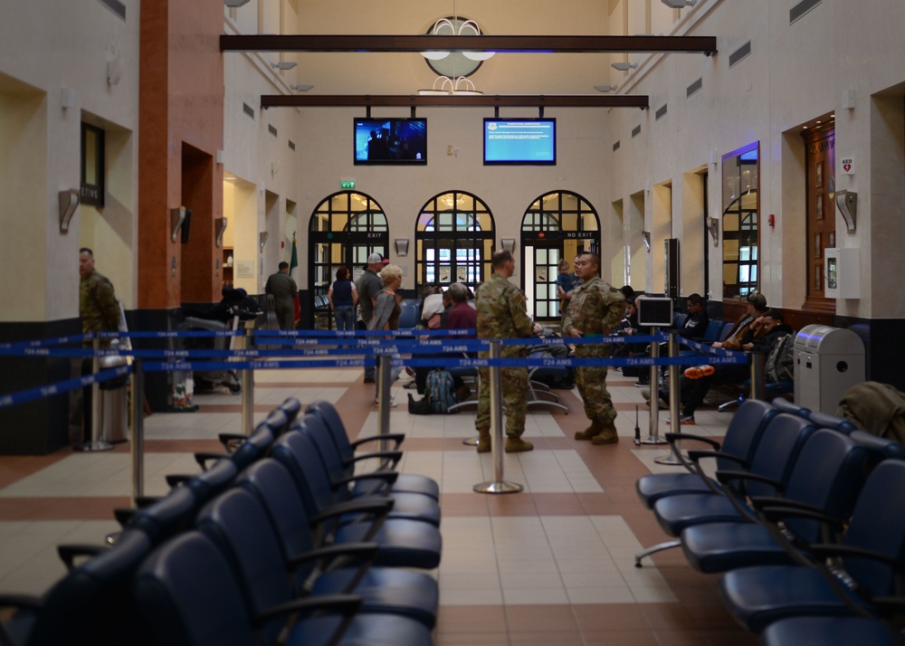 The image shows a military passenger terminal waiting area with blue queue barriers labeled '724 AMS' (Air Mobility Squadron). Several military personnel in camouflage uniforms and civilians are present in the terminal, which features arched windows, tiled flooring, and overhead monitors displaying information. The facility resembles a military air terminal used for Space-A (Space Available) or official military travel.