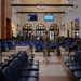 The image shows a military passenger terminal waiting area with blue queue barriers labeled '724 AMS' (Air Mobility Squadron). Several military personnel in camouflage uniforms and civilians are present in the terminal, which features arched windows, tiled flooring, and overhead monitors displaying information. The facility resembles a military air terminal used for Space-A (Space Available) or official military travel.
