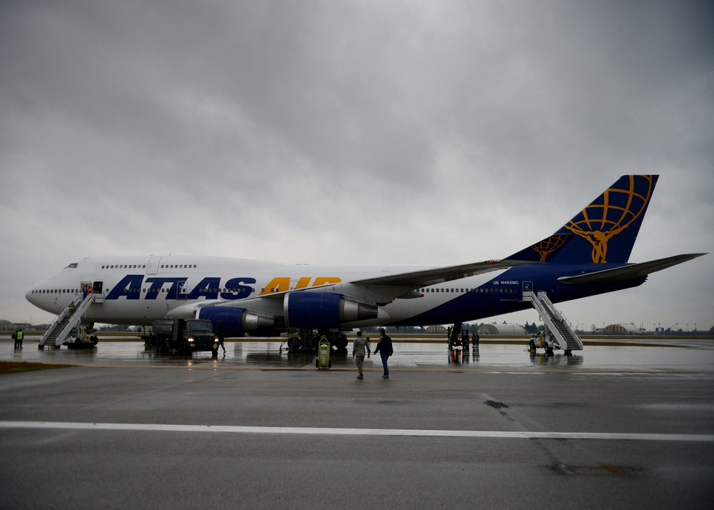 An Atlas Air Boeing 747 wide-body aircraft is parked on a wet tarmac under overcast, stormy skies. The aircraft displays the distinctive Atlas Air livery with a white upper fuselage, dark blue lower section, and gold globe-and-Atlas logo on the tail. Ground personnel and passengers are visible near the aircraft, with boarding stairs deployed at both the front and rear doors.