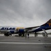 An Atlas Air Boeing 747 wide-body aircraft is parked on a wet tarmac under overcast, stormy skies. The aircraft displays the distinctive Atlas Air livery with a white upper fuselage, dark blue lower section, and gold globe-and-Atlas logo on the tail. Ground personnel and passengers are visible near the aircraft, with boarding stairs deployed at both the front and rear doors.