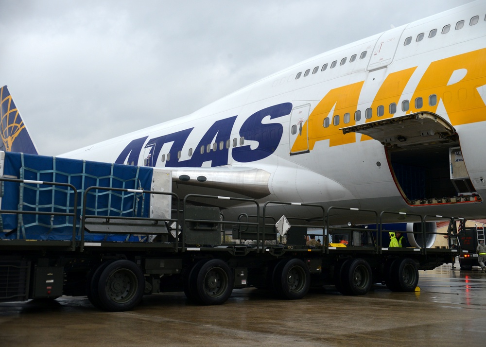 A large Atlas Air cargo Boeing 747 aircraft is shown on a wet airport tarmac during overcast conditions. Ground crew are actively loading or unloading cargo through the open main deck cargo door on the forward fuselage. Military-style flatbed cargo trailers loaded with blue tarp-covered freight are positioned alongside the aircraft.