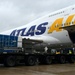 A large Atlas Air cargo Boeing 747 aircraft is shown on a wet airport tarmac during overcast conditions. Ground crew are actively loading or unloading cargo through the open main deck cargo door on the forward fuselage. Military-style flatbed cargo trailers loaded with blue tarp-covered freight are positioned alongside the aircraft.