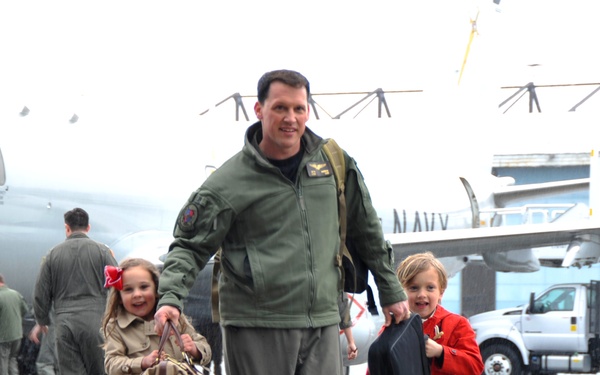 A U.S. Navy aviator in an olive green flight suit walks across a wet tarmac with two young children, appearing to return from a deployment. The man carries a large canvas duffel bag while the children assist with luggage, all three smiling. A U.S. Navy aircraft is visible in the background along with other flight crew members.