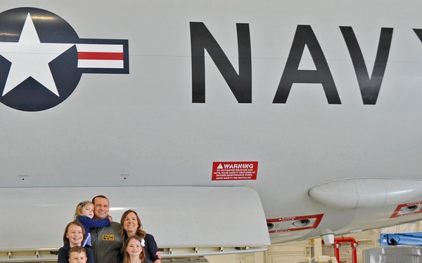 A family of six poses for a photograph inside a military hangar beneath a large U.S. Navy aircraft. The group includes one adult male in an olive green flight suit, one adult female in civilian clothing, and four children dressed in coordinating navy blue outfits. The setting is a well-lit, clean maintenance hangar with ground support equipment visible in the background.