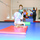 A gymnastics class is taking place in an indoor gym facility. A young blonde child is performing a forward roll or cartwheel on a red mat in the foreground, while an older girl in a black and teal leotard stands behind her. An instructor with arms raised and another child are visible in the background near a large mirror.